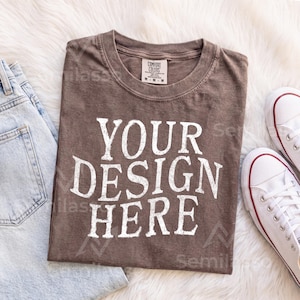 May include: A brown short-sleeved t-shirt with a crew neckline and the white text "YOUR DESIGN HERE" printed in a bold, distressed font on the front. The shirt features a size M tag on the inner collar and has a visible, textured fabric finish.