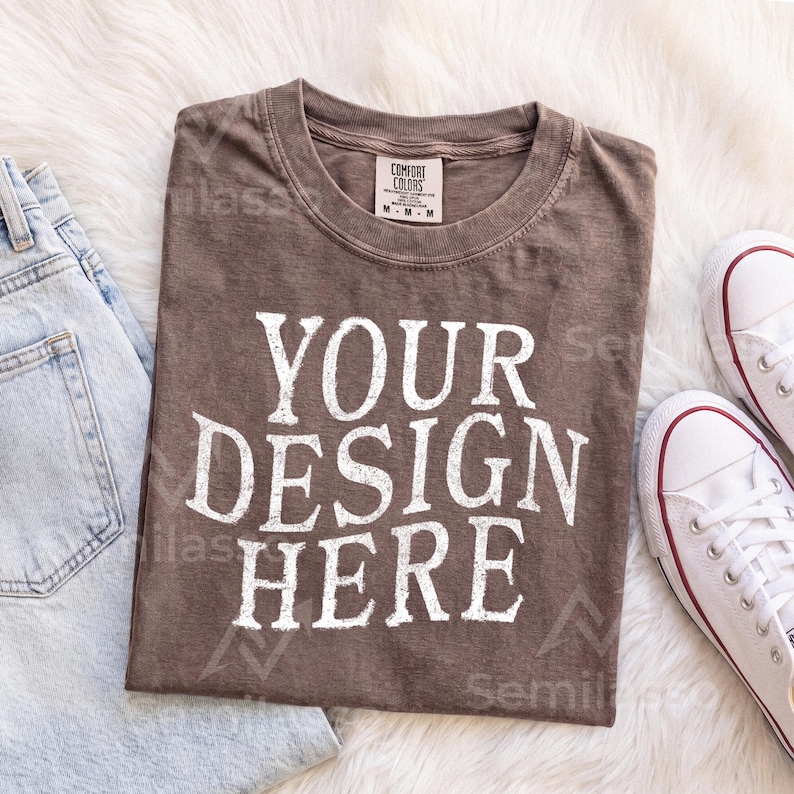 May include: A brown short-sleeved t-shirt with a crew neckline and the white text "YOUR DESIGN HERE" printed in a bold, distressed font on the front. The shirt features a size M tag on the inner collar and has a visible, textured fabric finish.