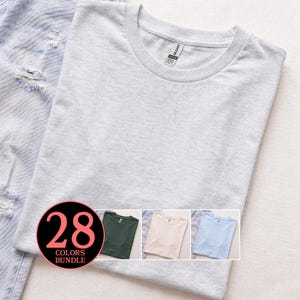 Puede incluir: Una camiseta gris claro con cuello redondo es el punto focal de esta imagen, con una etiqueta visible en el cuello. La camiseta está doblada cuidadosamente sobre un par de jeans azul claro. Un gráfico circular muestra "28 COLORS BUNDLE" con opciones de color adicionales.