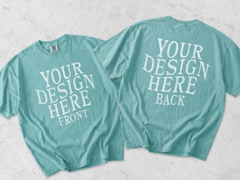 May include: Two teal short-sleeve t-shirts displayed together. The left shirt features white text reading YOUR DESIGN HERE FRONT. The right shirt displays white text reading YOUR DESIGN HERE BACK. Both shirts have a crew neckline and a relaxed fit.