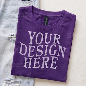 May include: A purple crew neck t-shirt with the text "YOUR DESIGN HERE" printed in a distressed white font on the front. The shirt features a black Gildan brand neck label and is positioned next to a pair of light blue distressed denim jeans.
