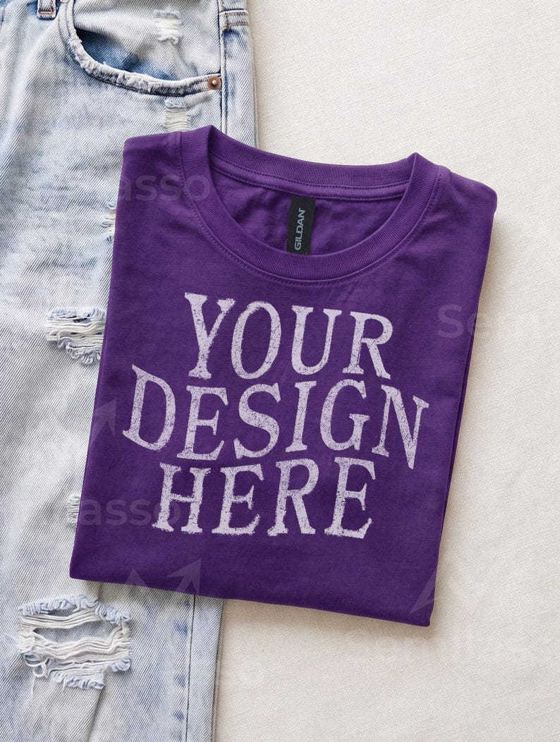 May include: A purple crew neck t-shirt with the text "YOUR DESIGN HERE" printed in a distressed white font on the front. The shirt features a black Gildan brand neck label and is positioned next to a pair of light blue distressed denim jeans.