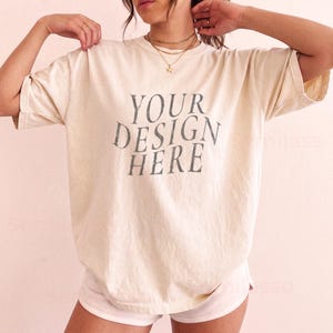 Puede incluir: Una camiseta de corte oversize en color crema con el texto "YOUR DESIGN HERE" impreso en un tono gris carbón oscuro y desgastado en el pecho. La prenda cuenta con mangas cortas y cuello redondo, ofreciendo un ajuste holgado y cómodo.