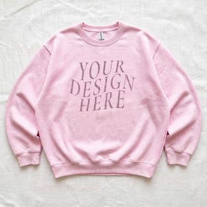 Puede incluir: Una sudadera de cuello redondo en color rosa claro con mangas largas y bordes acanalados en el cuello, pu&ntilde;os y dobladillo. En el centro del pecho aparece el texto "YOUR DESIGN HERE" en una tipograf&iacute;a serif de color rosa oscuro desgastada.