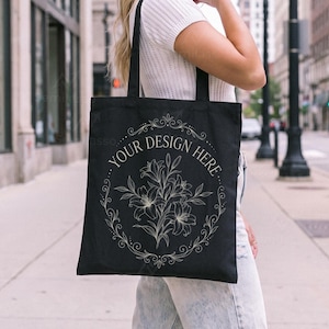 May include: A black tote bag featuring a white line art illustration of a lily bouquet enclosed within an ornate circular frame. The text "YOUR DESIGN HERE" is printed in a curved arc along the top portion of the decorative floral border.
