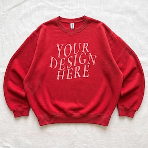 Puede incluir: Una sudadera roja de cuello redondo con mangas largas y pu&ntilde;os acanalados. En el centro del pecho aparece el texto blanco "YOUR DESIGN HERE" en una tipograf&iacute;a serif desgastada. La prenda incluye una etiqueta interior en el cuello.