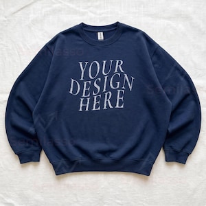 Puede incluir: Una sudadera azul marino de cuello redondo con el texto blanco "YOUR DESIGN HERE" impreso en una fuente serif desgastada en el pecho. La prenda tiene mangas largas, cuello y pu&ntilde;os acanalados, con una etiqueta de la marca Gildan en el cuello.