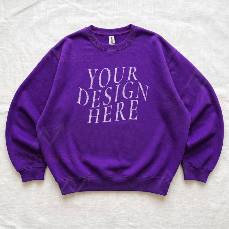 Puede incluir: Una sudadera de color morado vibrante con cuello redondo y manga larga, que presenta el texto "YOUR DESIGN HERE" impreso en una fuente blanca desgastada en el pecho. La prenda tiene cuello, pu&ntilde;os y dobladillo acanalados y una etiqueta Gildan.