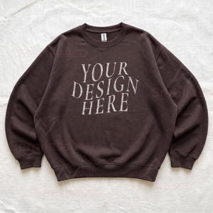 Puede incluir: Una sudadera de cuello redondo en color marr&oacute;n oscuro con el texto "YOUR DESIGN HERE" impreso en una fuente desgastada de color blanco roto en el pecho. La prenda tiene mangas largas, cuello y pu&ntilde;os acanalados, con una etiqueta Gildan en el cuello.