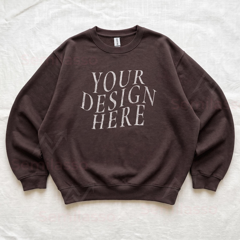 Puede incluir: Una sudadera de cuello redondo en color marr&oacute;n oscuro con el texto "YOUR DESIGN HERE" impreso en una fuente desgastada de color blanco roto en el pecho. La prenda tiene mangas largas, cuello y pu&ntilde;os acanalados, con una etiqueta Gildan en el cuello.