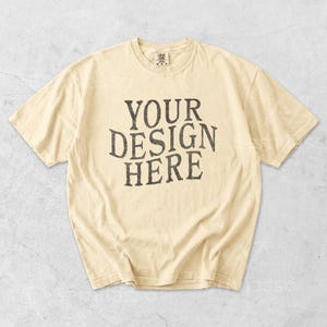 May include: Cream-colored t-shirt with the text "YOUR DESIGN HERE" in a distressed, dark gray font. The shirt has short sleeves and a crew neck. The fabric appears soft and slightly worn.