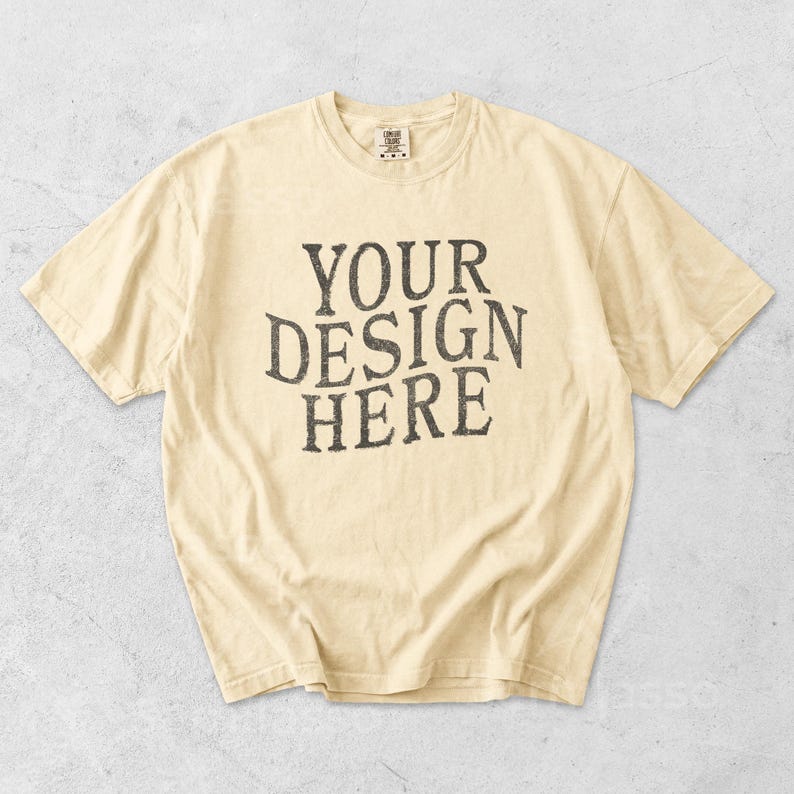 May include: Cream-colored t-shirt with the text "YOUR DESIGN HERE" in a distressed, dark gray font. The shirt has short sleeves and a crew neck. The fabric appears soft and slightly worn.