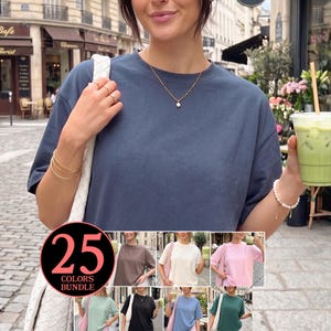 Puede incluir: Una camiseta azul marino de manga corta. La imagen muestra a una persona que lleva la camiseta, con un bolso blanco sobre el hombro y sosteniendo una bebida verde. Un gráfico en la parte inferior muestra la camiseta en varios colores, con el texto "25 COLORS BUNDLE".