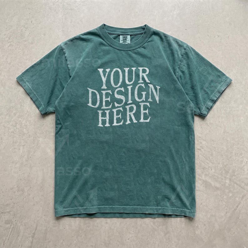 May include: A short-sleeve crew neck t-shirt in a muted teal green color with a vintage-style wash. The center of the chest features the white text "YOUR DESIGN HERE" in a bold, serif font. The garment has a standard fit and a ribbed collar.