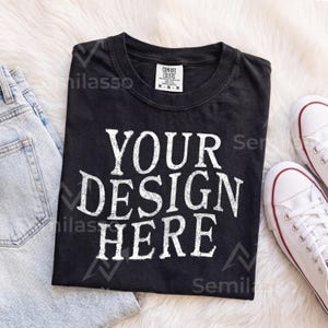 May include: A black short-sleeve t-shirt with a crew neckline featuring the white distressed-style text "YOUR DESIGN HERE" printed on the front. The shirt has a Comfort Colors brand label on the inside collar indicating a size medium.