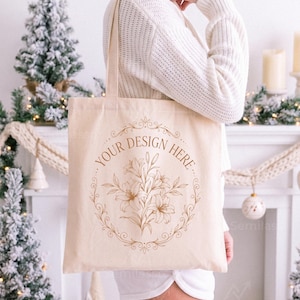 May include: A beige canvas tote bag featuring a brown line art illustration of a lily bouquet enclosed within an ornate circular frame. The text "YOUR DESIGN HERE" is printed in a curved brown font along the top of the decorative floral border.