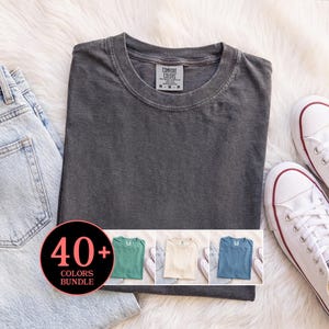 May include: A charcoal gray crew neck t-shirt with a Comfort Colors label, accompanied by light blue denim jeans and white canvas sneakers. A circular graphic overlay displays the text 40+ COLORS BUNDLE with smaller t-shirt images.
