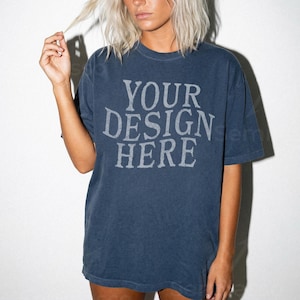 Puede incluir: Una camiseta azul marino con el texto "YOUR DESIGN HERE" en una fuente gris claro descolorida. La camiseta tiene mangas cortas y un corte holgado. La imagen muestra la camiseta en una persona.