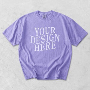 May include: A lavender-colored t-shirt with the text "YOUR DESIGN HERE" in white block letters. The short-sleeved shirt is made of a soft material and has a crew neck. The shirt is laid flat on a light gray surface.