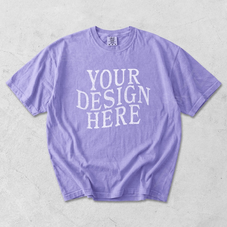 May include: A lavender-colored t-shirt with the text "YOUR DESIGN HERE" in white block letters. The short-sleeved shirt is made of a soft material and has a crew neck. The shirt is laid flat on a light gray surface.