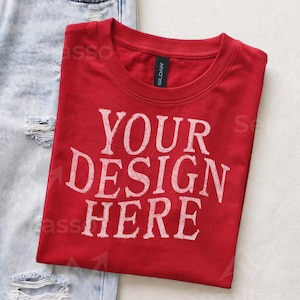 May include: A red short-sleeved t-shirt with a crew neckline and the white distressed text "YOUR DESIGN HERE" printed on the front. The shirt features a black Gildan brand label at the inner neck and is positioned next to light blue distressed denim.