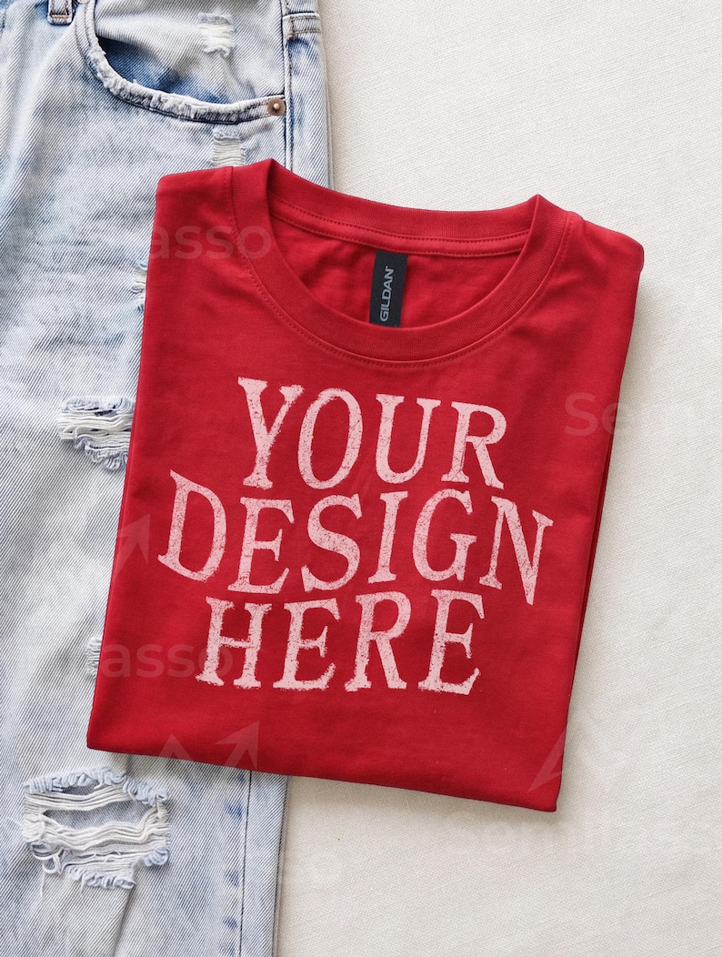 May include: A red short-sleeved t-shirt with a crew neckline and the white distressed text "YOUR DESIGN HERE" printed on the front. The shirt features a black Gildan brand label at the inner neck and is positioned next to light blue distressed denim.