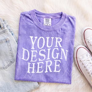May include: A purple short-sleeved t-shirt with a crew neckline and a white distressed graphic print that reads YOUR DESIGN HERE. The shirt features a Comfort Colors brand label on the inside collar and has a faded, vintage-style fabric texture.