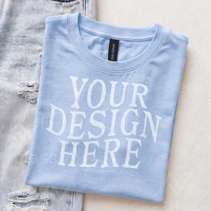 May include: A light blue short-sleeved t-shirt with a crew neckline and the white text "YOUR DESIGN HERE" printed on the front. The shirt features a black Gildan brand label at the inner neck and is positioned next to a pair of light blue distressed denim jeans.