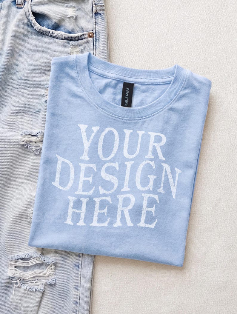 May include: A light blue short-sleeved t-shirt with a crew neckline and the white text "YOUR DESIGN HERE" printed on the front. The shirt features a black Gildan brand label at the inner neck and is positioned next to a pair of light blue distressed denim jeans.