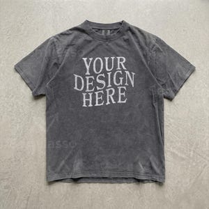 May include: A charcoal grey short-sleeve t-shirt with a crew neckline. The front of the shirt features the white text "YOUR DESIGN HERE" printed in a bold, sans-serif font. The fabric has a washed, slightly faded appearance throughout the garment.