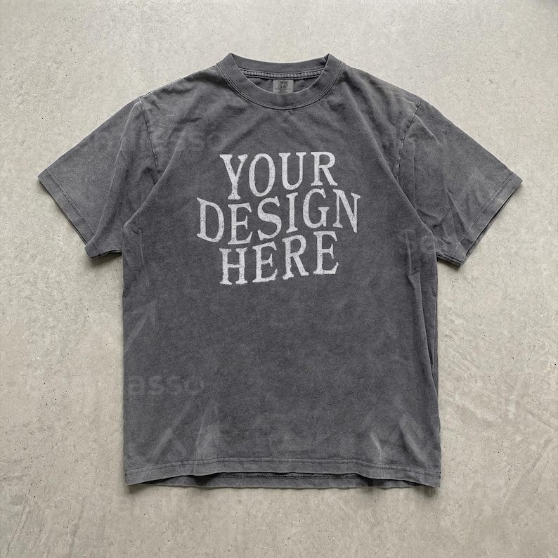 May include: A charcoal grey short-sleeve t-shirt with a crew neckline. The front of the shirt features the white text "YOUR DESIGN HERE" printed in a bold, sans-serif font. The fabric has a washed, slightly faded appearance throughout the garment.