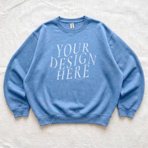 Puede incluir: Una sudadera de cuello redondo en color azul claro con el texto blanco "YOUR DESIGN HERE" impreso en una fuente serif en el pecho. La prenda tiene mangas largas, cuello, pu&ntilde;os y dobladillo acanalados, con una peque&ntilde;a etiqueta blanca en el cuello.