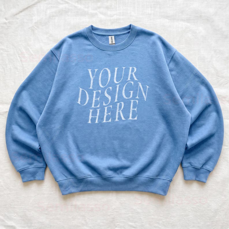 Puede incluir: Una sudadera de cuello redondo en color azul claro con el texto blanco "YOUR DESIGN HERE" impreso en una fuente serif en el pecho. La prenda tiene mangas largas, cuello, pu&ntilde;os y dobladillo acanalados, con una peque&ntilde;a etiqueta blanca en el cuello.