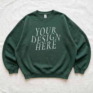 Puede incluir: Una sudadera de cuello redondo en color verde bosque oscuro con el texto blanco "YOUR DESIGN HERE" impreso en una fuente serif desgastada en el pecho. La prenda tiene mangas largas, cuello acanalado y pu&ntilde;os acanalados, con una etiqueta Gildan.