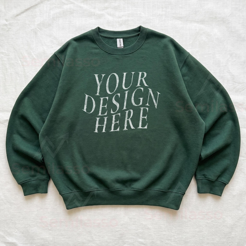 Puede incluir: Una sudadera de cuello redondo en color verde bosque oscuro con el texto blanco "YOUR DESIGN HERE" impreso en una fuente serif desgastada en el pecho. La prenda tiene mangas largas, cuello acanalado y pu&ntilde;os acanalados, con una etiqueta Gildan.