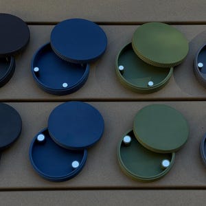 Può includere: Otto piccoli contenitori rotondi con finitura opaca in vari colori: nero, blu navy, verde oliva e grigio. Ogni contenitore ha un coperchio abbinato e un piccolo magnete argentato all'interno. Su una superficie di legno marrone.
