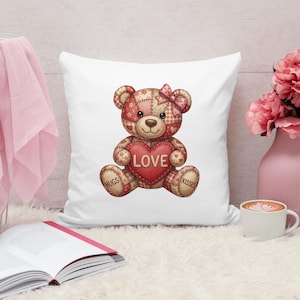 May include: White square cushion featuring a patchwork teddy bear holding a red heart with the word "LOVE". The bear has a pink bow and the words "HUGS" and "KISSES" on its paws. The cushion is on a white fluffy surface.