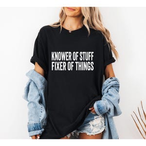 Puede incluir: Camiseta negra con el texto blanco "KNOWER OF STUFF FIXER OF THINGS". La camiseta se usa con una chaqueta vaquera azul claro y pantalones cortos vaqueros. El fondo es una pared blanca lisa.