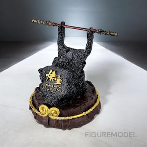 May include: A black statue with gold accents, featuring a hand holding a staff. The base is brown with gold trim, and the statue has the text "BLACK MYTH WUKONG" in gold. The staff is brown and red.