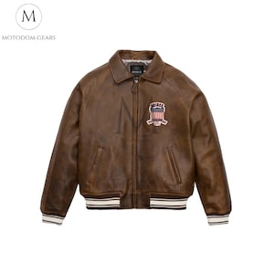May include: Brown leather bomber jacket with a zip closure, collar, and ribbed cuffs and hem. Features a patch with a shield design. The jacket has the brand name "AVIREX" on the zipper and the brand name "MOTODOM GEARS" on the upper left.