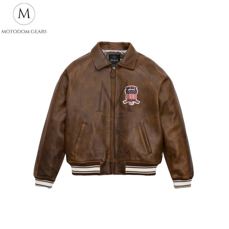 May include: Brown leather bomber jacket with a zip closure, collar, and ribbed cuffs and hem. Features a patch with a shield design. The jacket has the brand name "AVIREX" on the zipper and the brand name "MOTODOM GEARS" on the upper left.
