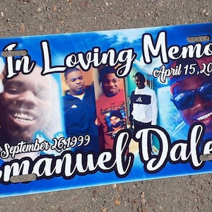 May include: A blue license plate with the words "In Loving Memory" and "Emanuel Dale" in white script. It features photos of four people and the dates "September 26, 1999" and "April 15, 2020". White doves are also present.