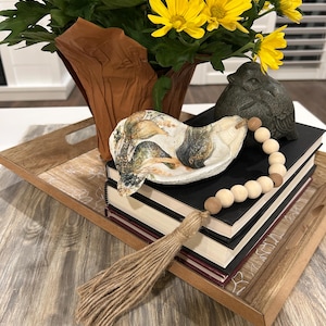May include: A decorative tray holds a stack of books, a seashell-shaped dish, a small bird statue, and a beaded garland with a tassel. A bouquet of yellow flowers in a brown paper-lined vase sits in the background. The tray has a wooden frame.