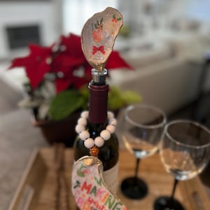 May include: A wine bottle with a decorative oyster shell bottle stopper featuring a cocktail design and a red bow. A second oyster shell with the text "MAKING spirits BRIGHT" hangs from the bottle. White beaded necklace and two wine glasses are also visible.
