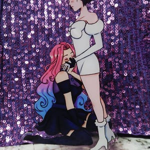 May include: An acrylic anime-style figure featuring two female characters. One is in a white dress and the other in a dark outfit with pink and blue hair. The figure is on a clear base, with a purple sequined background.