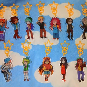May include: A selection of character keyrings. Each keyring displays a vibrant anime-style illustration of a person. The keyrings are connected to gold-coloured star-shaped clasps. The background is a light blue fabric with a cloud pattern.
