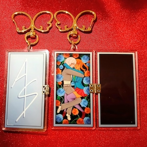 May include: Two gold keyrings with clear acrylic panels. One panel has a light blue background with white script. The other panel features a couple embracing, surrounded by blue and orange roses.
