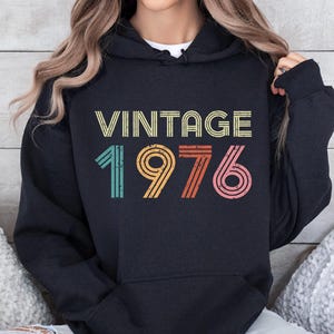 May include: A black hooded sweatshirt featuring the text VINTAGE 1976 in a distressed, multi-colored font. The design includes horizontal lines in shades of yellow, teal, orange, and pink, with a front kangaroo pocket and long sleeves.