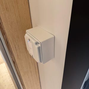 May include: A white electrical box mounted on a white wall next to a wooden door frame. The box has a small, rectangular design with a visible switch and a blue indicator light. The box is secured with screws.