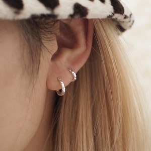 May include: Two small, silver-toned hoop earrings with a hammered, textured finish. The earrings are worn in the earlobe and the upper cartilage, featuring a reflective metallic surface and a circular, continuous design that catches the light.